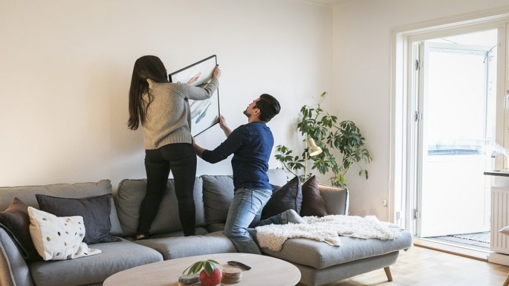 Couple Adjusting Painting On Wall While Leaning On Sofa At Home