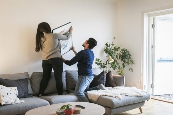 Couple Adjusting Painting On Wall While Leaning On Sofa At Home